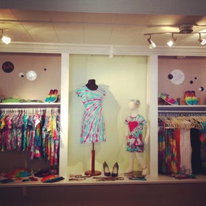 May include: A boutique display featuring tie-dye clothing. A dress and a child's dress are displayed on mannequins. Racks of colorful tie-dye shirts and accessories are visible. Shoes and accessories are on display shelves. The word "Kids" is on a sign.