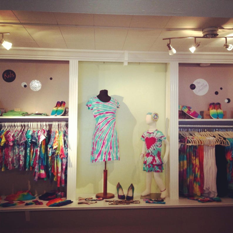 May include: A boutique display featuring tie-dye clothing. A dress and a child's dress are displayed on mannequins. Racks of colorful tie-dye shirts and accessories are visible. Shoes and accessories are on display shelves. The word "Kids" is on a sign.
