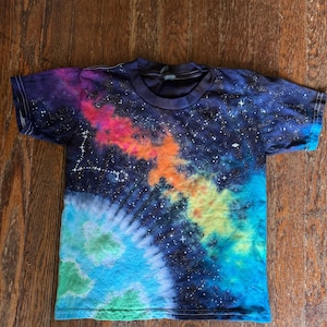 May include: A short-sleeve t-shirt featuring a vibrant galaxy print with deep blue, purple, pink, orange, yellow, and teal colors. The design includes scattered white star patterns and a constellation formation against a dark background.