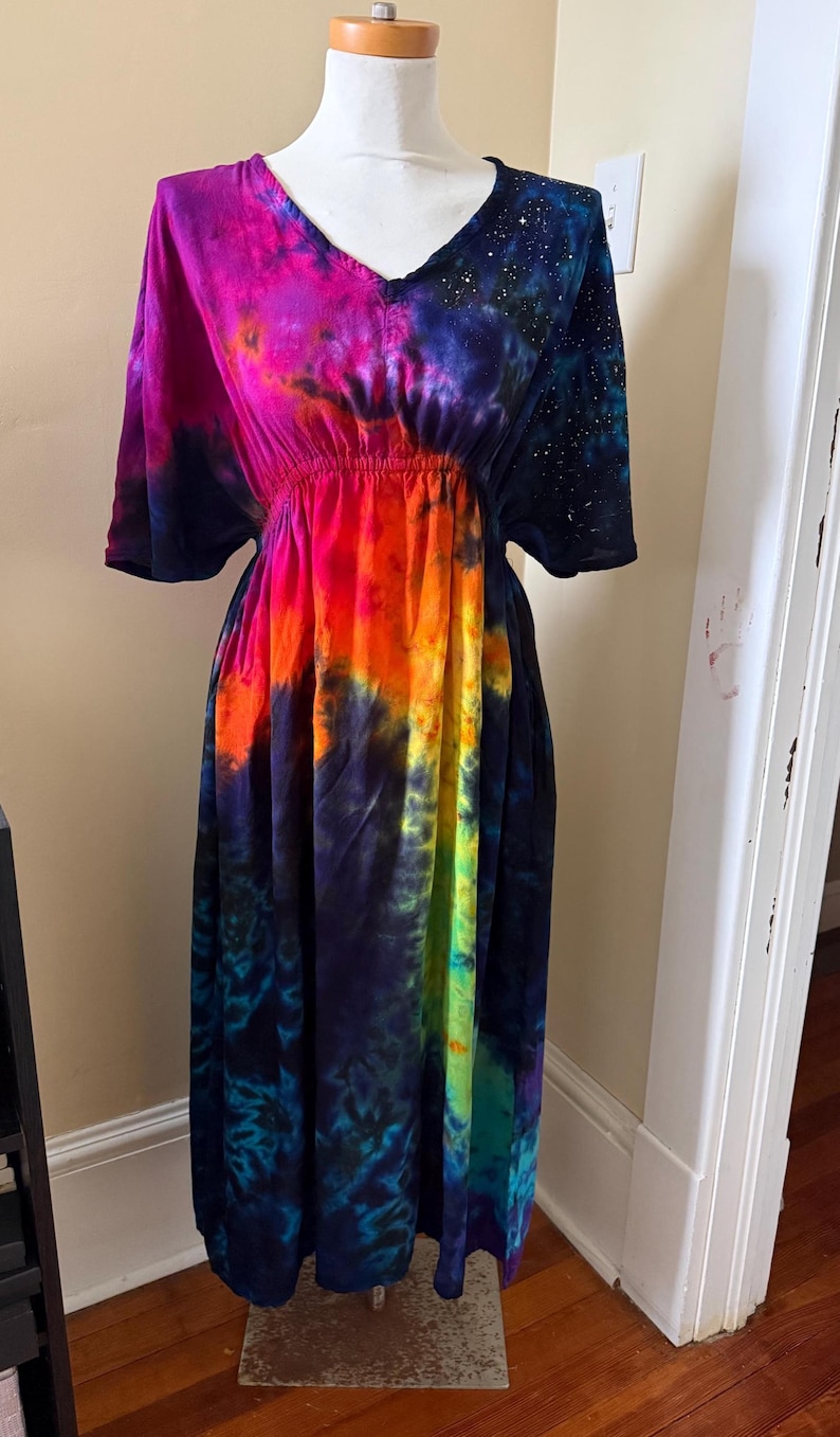 May include: A midi-length dress featuring a vibrant tie-dye pattern with shades of magenta, orange, yellow, and deep blue. The garment has short sleeves, a V-neckline, and an elasticized waist, with a galaxy-inspired print on the left shoulder.