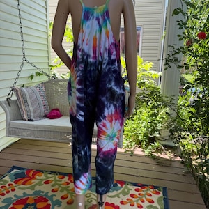 May include: A tie-dye jumpsuit in a variety of colors, including blue, purple, orange, and white, is displayed on a mannequin. The jumpsuit has a halter top and wide legs. The background includes a porch, a swing, and a colorful rug.
