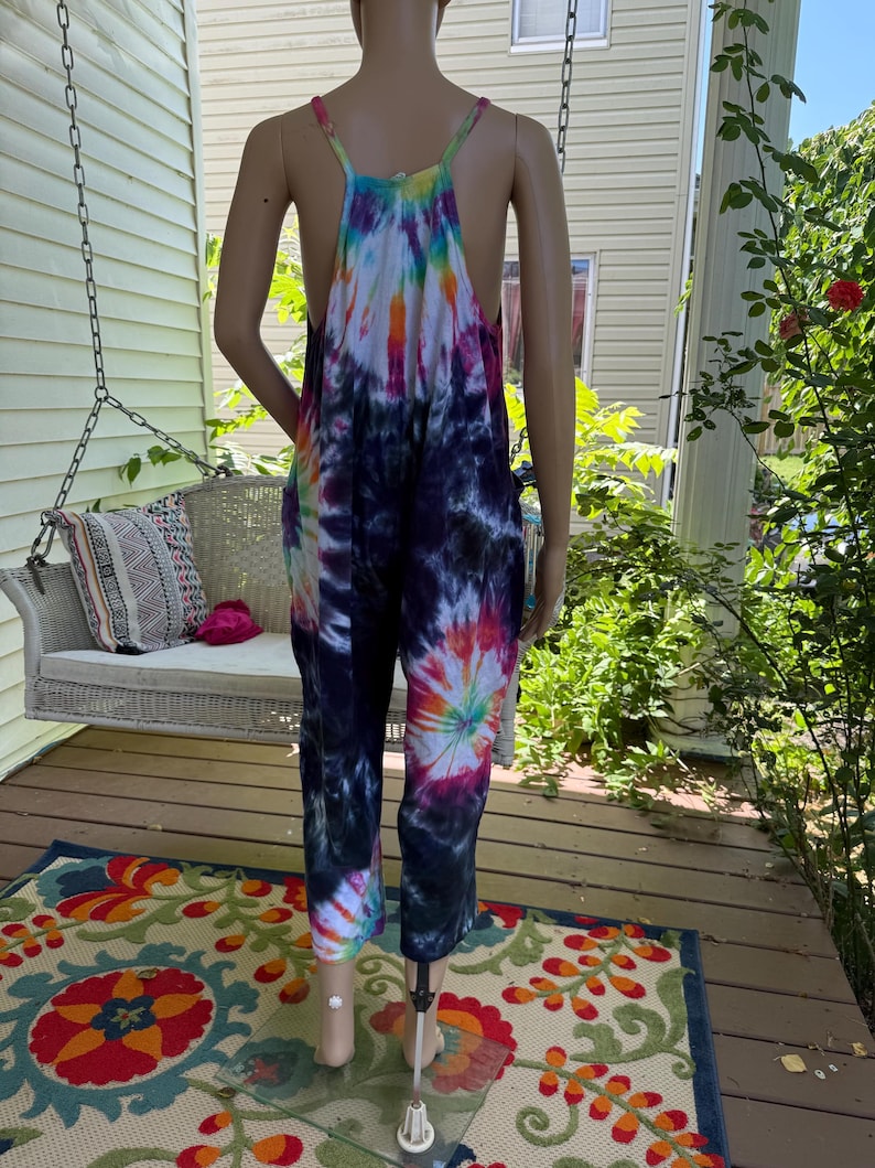 May include: A tie-dye jumpsuit in a variety of colors, including blue, purple, orange, and white, is displayed on a mannequin. The jumpsuit has a halter top and wide legs. The background includes a porch, a swing, and a colorful rug.