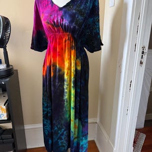 May include: A midi-length dress featuring a vibrant tie-dye pattern in shades of magenta, orange, yellow, and blue. The garment has short sleeves, a V-neckline, and an elasticized waist, with a dark blue section on the shoulder featuring small white dots.