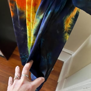 May include: A garment featuring a vibrant tie-dye pattern with bold streaks of orange, yellow, and deep blue. The fabric displays a swirling, blended color effect throughout the material, and the side of the piece includes a functional pocket opening.