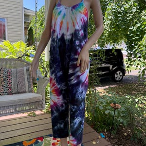 May include: A tie-dye jumpsuit in a variety of colors, including blue, purple, pink, and yellow. The jumpsuit has a scoop neckline, spaghetti straps, and cropped legs. The garment is displayed on a mannequin on a wooden porch.