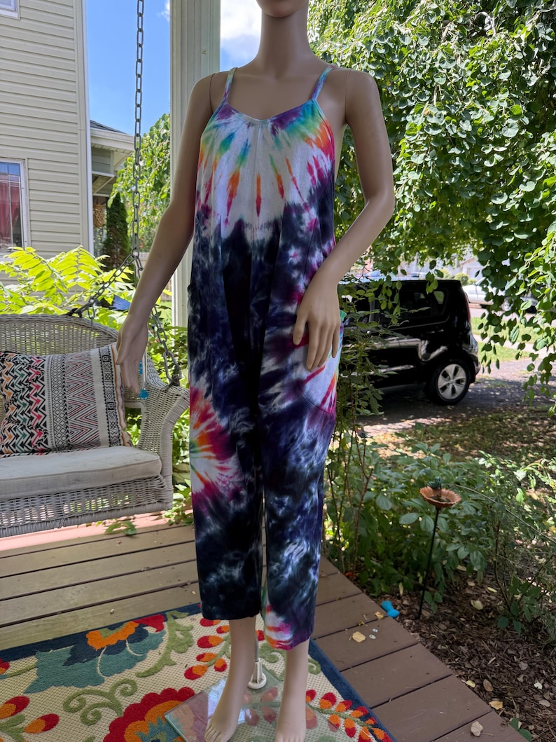 May include: A tie-dye jumpsuit in a variety of colors, including blue, purple, pink, and yellow. The jumpsuit has a scoop neckline, spaghetti straps, and cropped legs. The garment is displayed on a mannequin on a wooden porch.