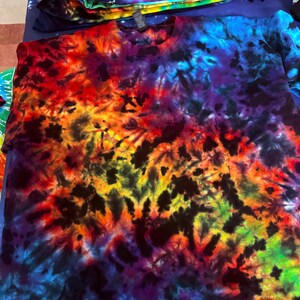 May include: A vibrant tie-dye t-shirt in a spectrum of colors, including red, orange, yellow, green, blue, and purple. The shirt has a crew neck and a unique, swirling pattern. Several other tie-dye shirts are stacked in the background.