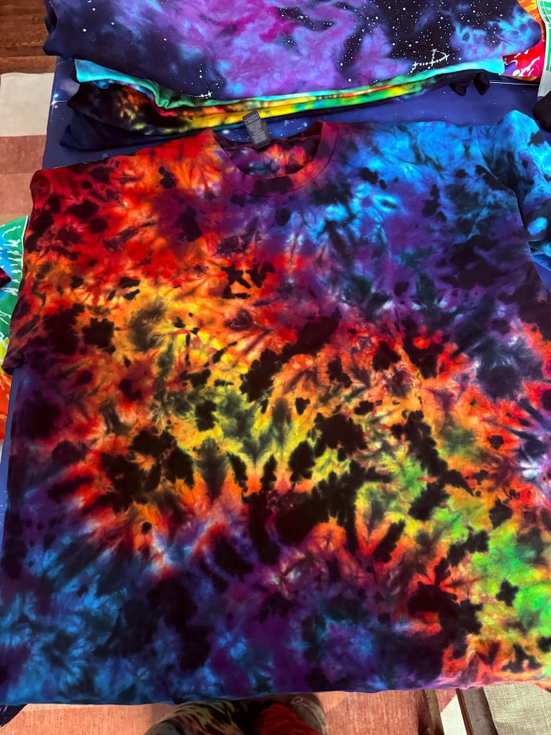 May include: A vibrant tie-dye t-shirt in a spectrum of colors, including red, orange, yellow, green, blue, and purple. The shirt has a crew neck and a unique, swirling pattern. Several other tie-dye shirts are stacked in the background.