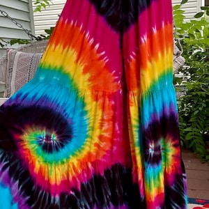 May include: A colourful, floor-length tie-dye skirt with a spiral design. The skirt displays a range of colours: pink, orange, yellow, green, blue, and purple, with black detailing. The skirt has a tiered design and a flowing shape.