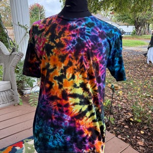 May include: A short-sleeved tie-dye t-shirt in a vibrant array of colors, including orange, yellow, pink, blue, and purple. The shirt has a black collar and is displayed on a mannequin. The tie-dye pattern is a swirling design.