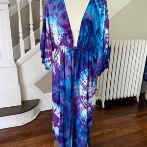 May include: A long, tie-dye kimono in shades of blue, purple, and white. The open-front robe features wide sleeves and a drawstring waist. The garment is displayed on a mannequin.