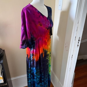 May include: A midi-length dress featuring a vibrant tie-dye pattern with bold swirls of magenta, orange, yellow, green, and deep blue. The garment has a V-neckline, short sleeves, and an elasticized waistline that creates a gathered, flowing skirt.
