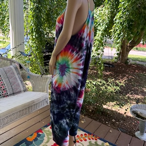 May include: A tie-dye jumpsuit in a vibrant spiral pattern of rainbow colors, including pink, orange, yellow, green, and purple, against a dark blue background. The jumpsuit has thin straps and wide legs, creating a relaxed, bohemian style.