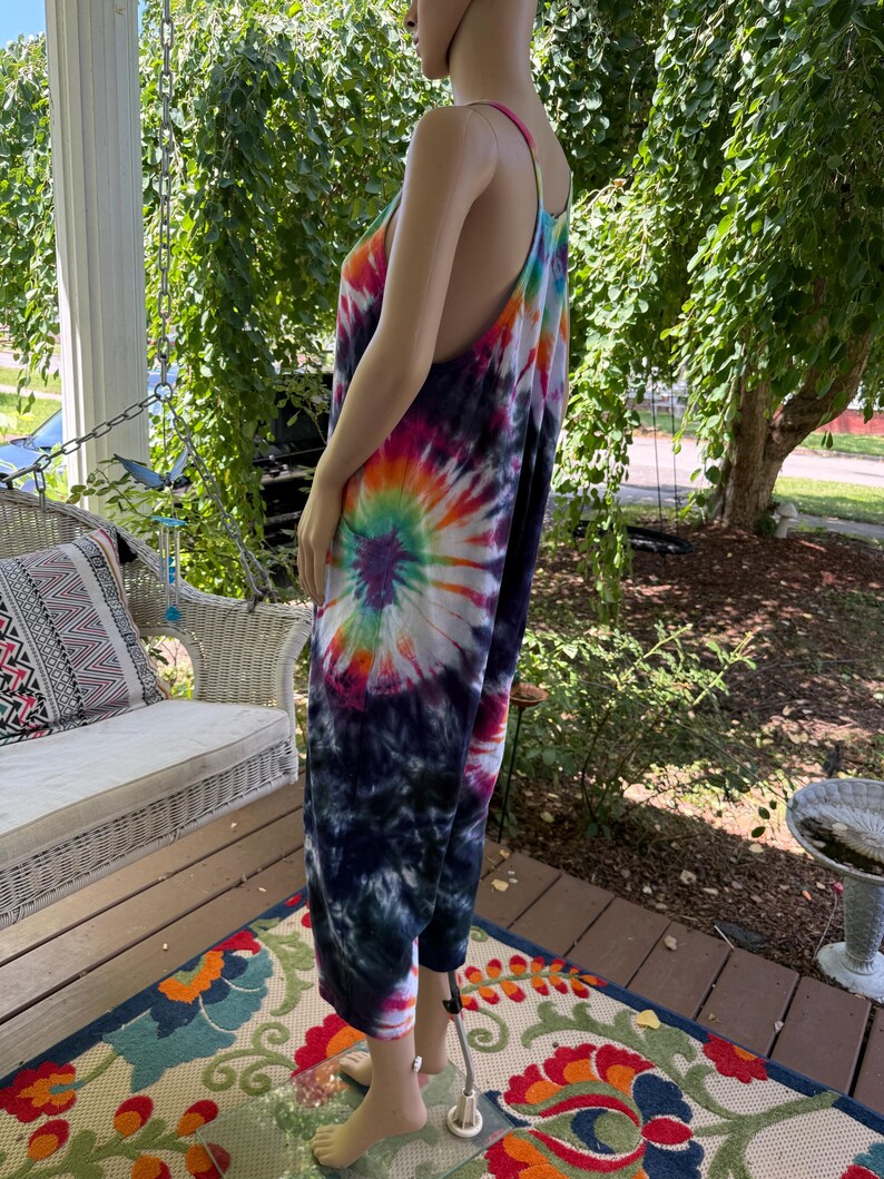 May include: A tie-dye jumpsuit in a vibrant spiral pattern of rainbow colors, including pink, orange, yellow, green, and purple, against a dark blue background. The jumpsuit has thin straps and wide legs, creating a relaxed, bohemian style.