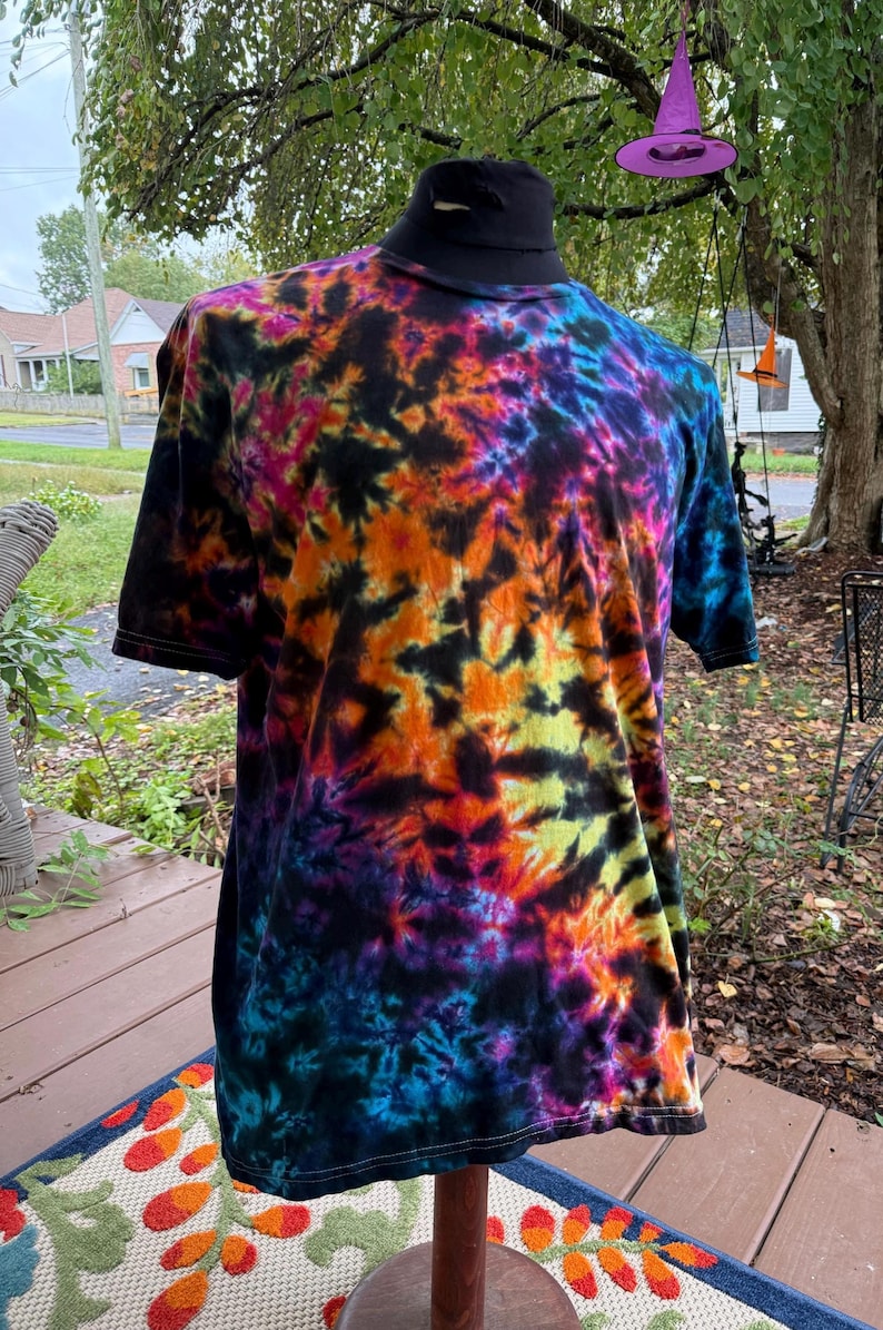 May include: A short-sleeved tie-dye t-shirt in a vibrant array of colors, including orange, yellow, blue, purple, and black. The shirt is displayed on a mannequin, showcasing its unique, colorful pattern. The shirt has a black collar.