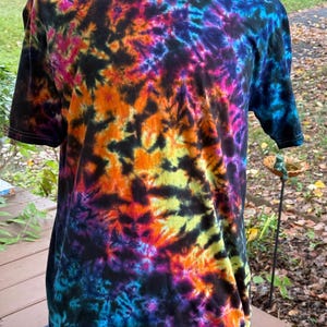 May include: A short-sleeved t-shirt with a vibrant tie-dye pattern. The shirt features a black base with swirling designs in shades of orange, yellow, pink, purple, and blue. The shirt is displayed on a mannequin.