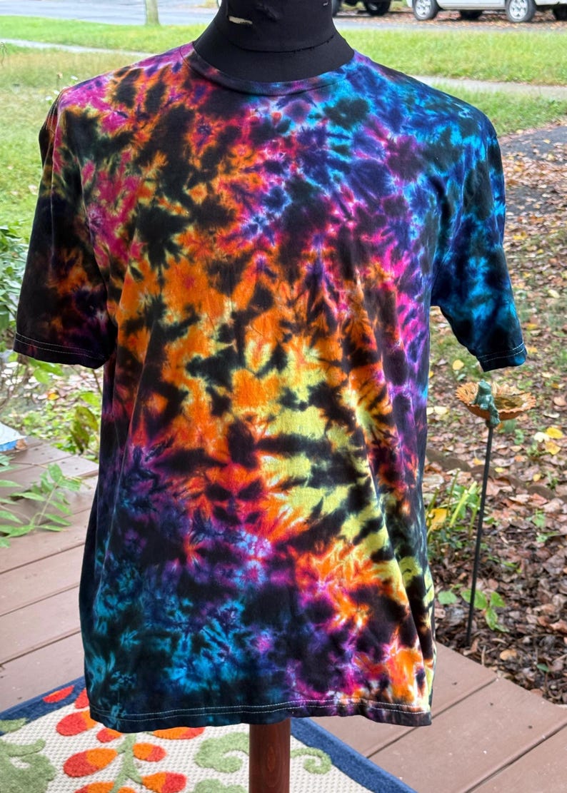 May include: A short-sleeved t-shirt with a vibrant tie-dye pattern. The shirt features a black base with swirling designs in shades of orange, yellow, pink, purple, and blue. The shirt is displayed on a mannequin.