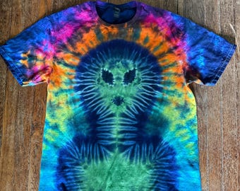 Pode incluir: Uma camiseta tie-dye com um design vibrante e psicodélico. A camisa apresenta uma figura central semelhante a um alienígena em tons de azul, verde e amarelo, cercada por um arco-íris de cores. A camisa está em uma superfície de madeira.