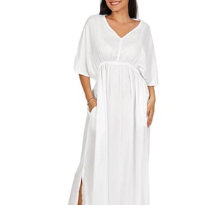 May include: A solid white maxi dress featuring a V-neckline, loose kimono-style sleeves, and an elasticized waist. The garment includes side pockets and a high side slit on the skirt, providing a relaxed fit and a flowing silhouette throughout.