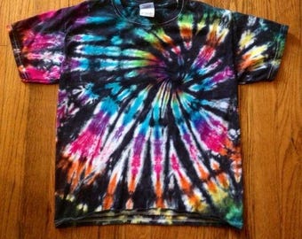 May include: A black tie-dye t-shirt with a spiral design in bright colours including pink, orange, yellow, green, blue, and purple. The shirt has a crew neck and short sleeves, and is displayed on a wooden surface.