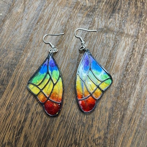 Pode incluir: Um par de brincos de asa de borboleta com cores do arco-íris e ganchos prateados. As asas são contornadas em preto e têm um efeito de vitral.