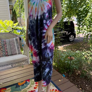 May include: A tie-dye jumpsuit in a vibrant array of colors, including blue, purple, pink, orange, and white. The jumpsuit features a halter neckline and cropped legs. The garment is displayed on a mannequin outdoors.