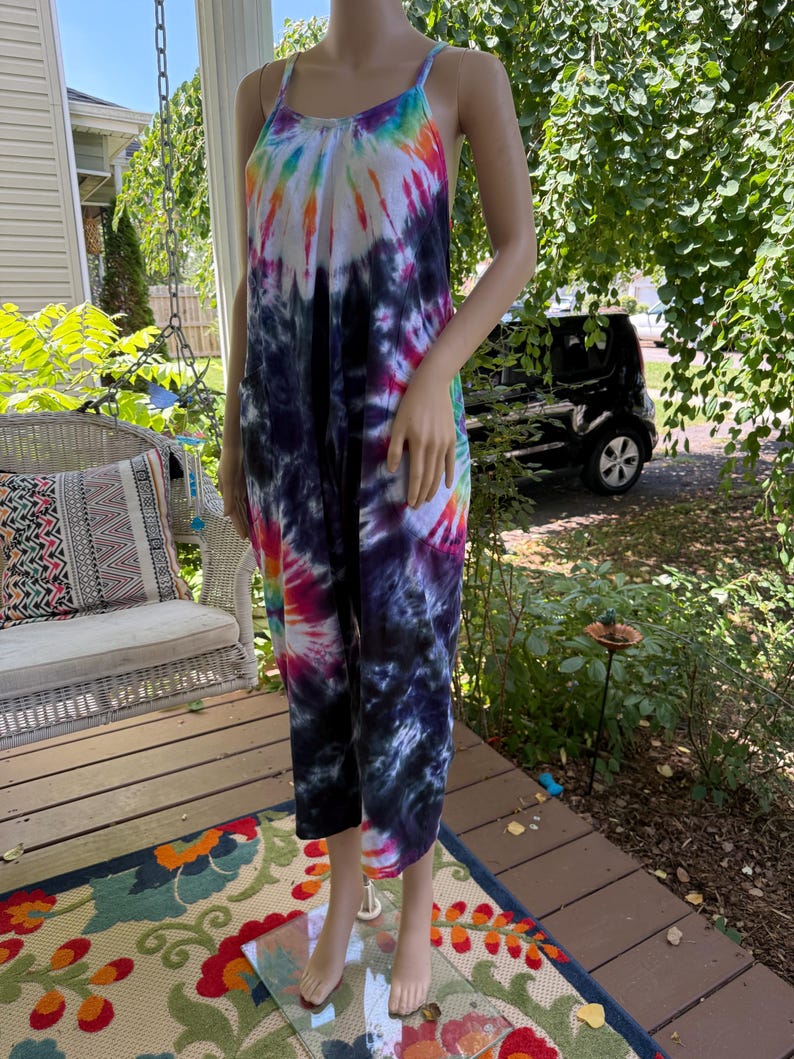May include: A tie-dye jumpsuit in a vibrant array of colors, including blue, purple, pink, orange, and white. The jumpsuit features a halter neckline and cropped legs. The garment is displayed on a mannequin outdoors.