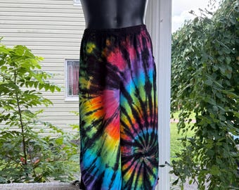 May include: Tie-dye pants in a vibrant spiral pattern of rainbow colors, including red, orange, yellow, green, blue, and purple. The pants are displayed on a black mannequin, showcasing the elastic waistband and loose fit. The background includes greenery and a white porch.