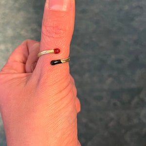 May include: A gold ring with a black and red enamel design. The ring is worn on a person's thumb.