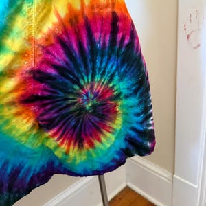 May include: A colourful tie-dye dress with a spiral design, showcasing a range of hues such as blue, green, yellow, orange, pink, and purple. The dress is presented on a metal stand, highlighting its unique pattern and style.