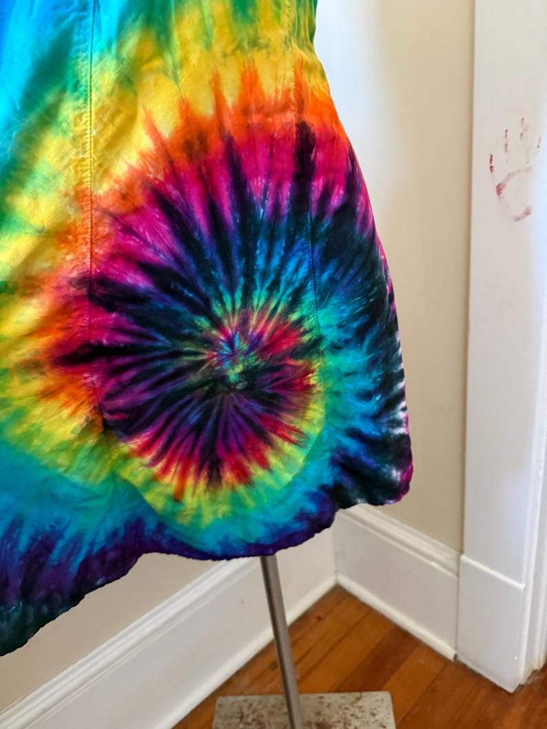 May include: A colourful tie-dye dress with a spiral design, showcasing a range of hues such as blue, green, yellow, orange, pink, and purple. The dress is presented on a metal stand, highlighting its unique pattern and style.