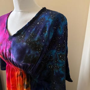 May include: A V-neck dress featuring a vibrant tie-dye pattern. The upper section displays a deep blue and black galaxy print with white star-like speckles, while the lower portion transitions into bright shades of pink, orange, and yellow.