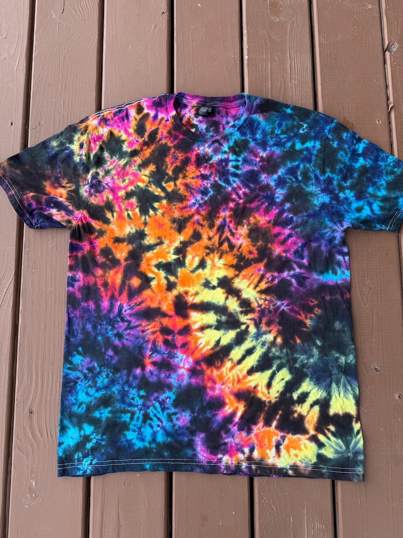 May include: A short-sleeved t-shirt with a vibrant tie-dye pattern. The shirt features a blend of colors, including black, blue, purple, pink, orange, and yellow. The tie-dye design is spread across the entire shirt, creating a psychedelic effect. The shirt is made of a soft material.