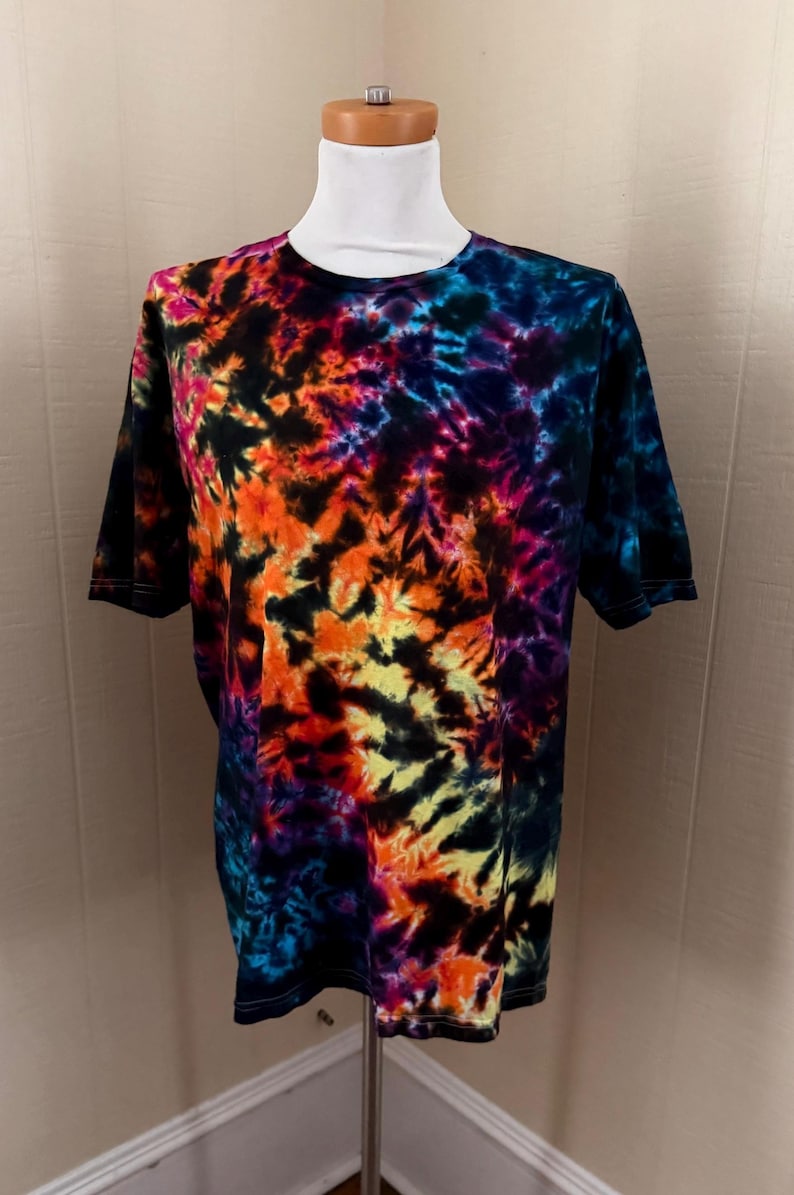 May include: A short-sleeved t-shirt with a vibrant tie-dye pattern. The shirt features a blend of colors, including orange, yellow, blue, purple, and black. The tie-dye design is displayed on a white mannequin.