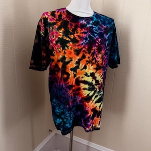 May include: A short-sleeved t-shirt with a vibrant tie-dye pattern. The shirt features a blend of colors, including black, orange, yellow, pink, and blue. The shirt is displayed on a mannequin.