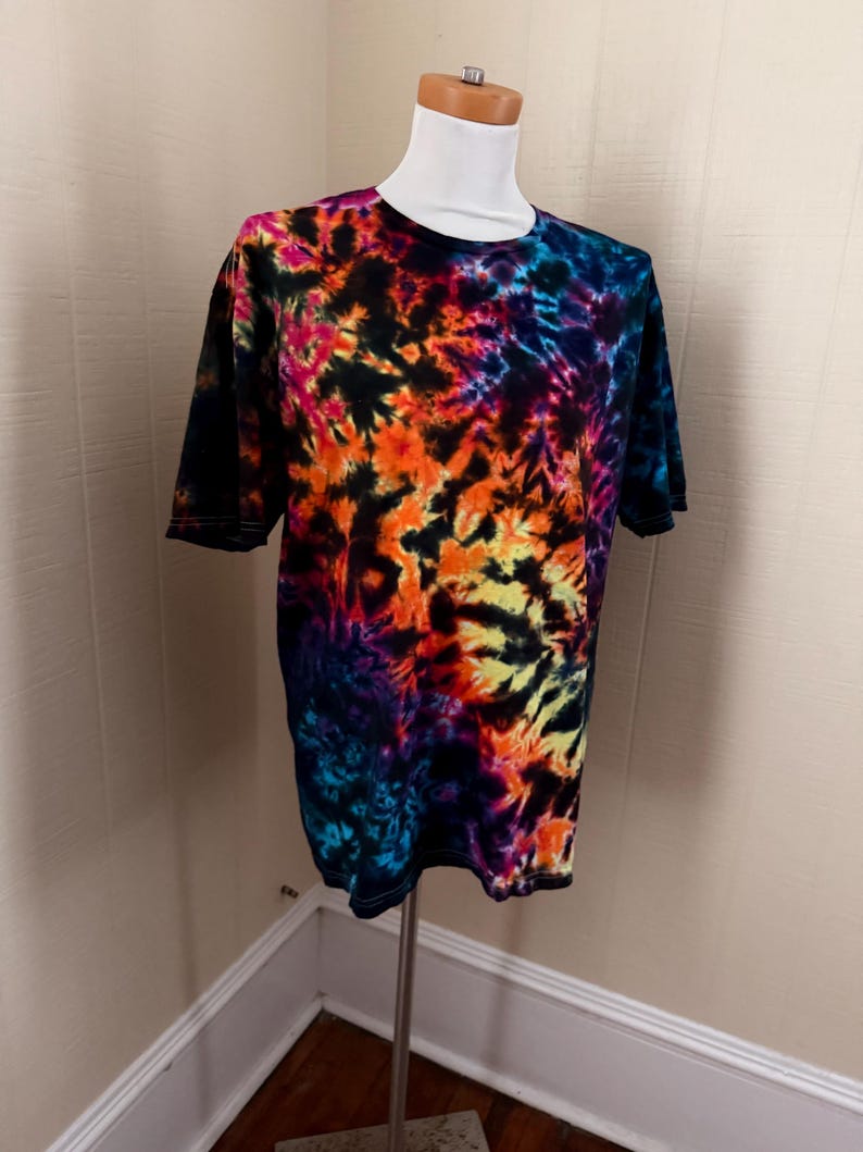 May include: A short-sleeved t-shirt with a vibrant tie-dye pattern. The shirt features a blend of colors, including black, orange, yellow, pink, and blue. The shirt is displayed on a mannequin.