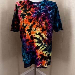May include: A short-sleeved t-shirt with a vibrant tie-dye pattern. The shirt features a blend of colors, including orange, yellow, blue, purple, and black. The shirt is displayed on a mannequin.