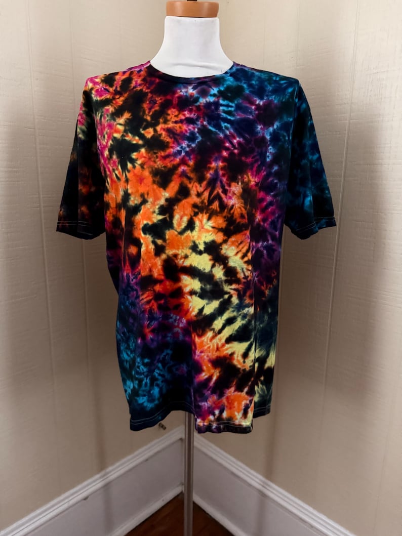 May include: A short-sleeved t-shirt with a vibrant tie-dye pattern. The shirt features a blend of colors, including orange, yellow, blue, purple, and black. The shirt is displayed on a mannequin.