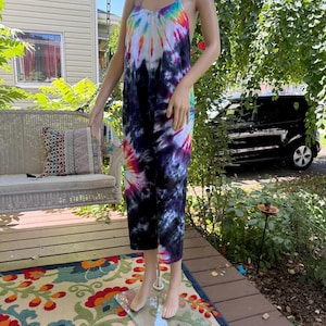 May include: A tie-dye jumpsuit with a scoop neck and spaghetti straps. The jumpsuit features a vibrant rainbow pattern at the top, transitioning to dark blue and purple hues. The model is standing on a glass stand on a porch.