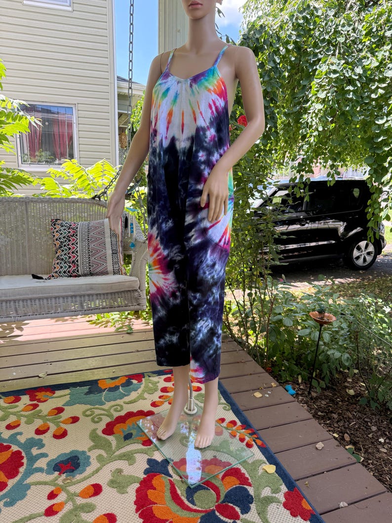May include: A tie-dye jumpsuit with a scoop neck and spaghetti straps. The jumpsuit features a vibrant rainbow pattern at the top, transitioning to dark blue and purple hues. The model is standing on a glass stand on a porch.