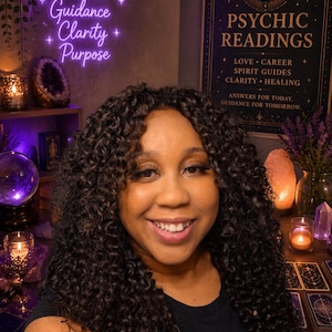 Peut inclure: Une femme aux cheveux bouclés foncés sourit devant une enseigne au néon qui dit "Intuition, Guidance, Clarity, Purpose". Une enseigne derrière elle dit "Psychic Readings" avec du texte sur l'amour, la carrière et la guérison. Des cartes de tarot et des cristaux sont visibles.