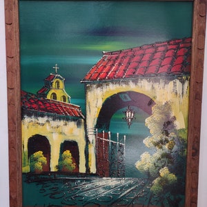 May include: A framed painting depicting a colorful scene of a building with a red tile roof, a green courtyard, and a small church with a cross on top. The painting is set against a dark green background and has a brown wooden frame.