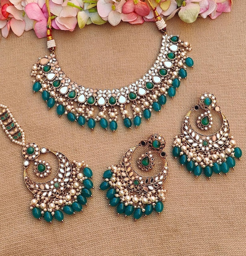 Puede incluir: Un collar, pendientes y un maang tikka de tono dorado con piedras verdes y blancas y detalles de perlas. Las joyas est&aacute;n dise&ntilde;adas en un estilo tradicional indio.