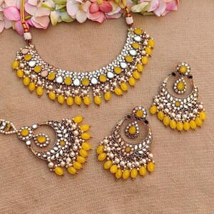 Puede incluir: Un conjunto de collar y pendientes amarillo y dorado con detalles de espejo y perlas. El collar tiene forma de l&aacute;grima y los pendientes son de forma de media luna.