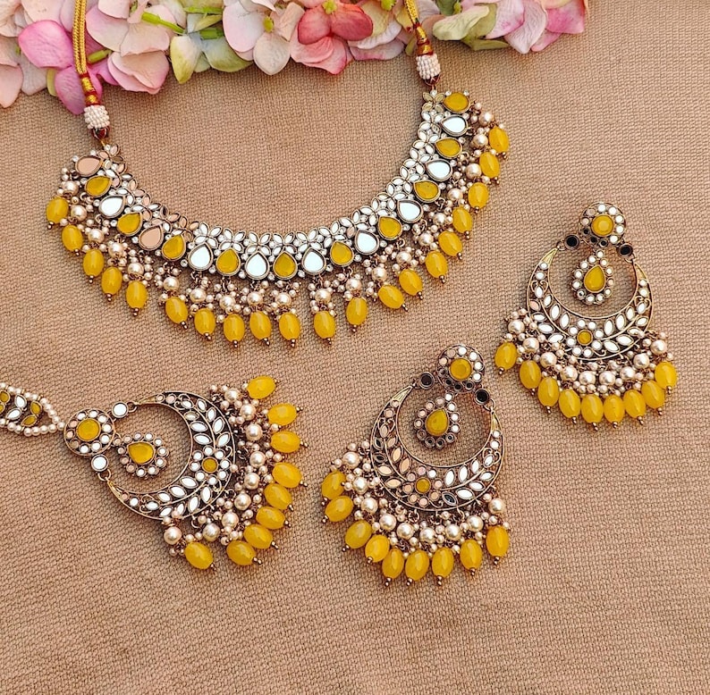 Puede incluir: Un conjunto de collar y pendientes amarillo y dorado con detalles de espejo y perlas. El collar tiene forma de l&aacute;grima y los pendientes son de forma de media luna.