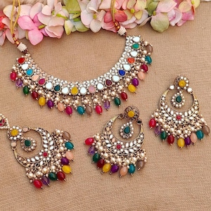 Puede incluir: Un conjunto de collar y pendientes de tono dorado con un dise&ntilde;o multicolor. El collar presenta un gran colgante redondo con una superficie de espejo y est&aacute; adornado con piedras de colores y perlas. Los pendientes tienen forma de media luna y presentan un dise&ntilde;o similar con piedras de colores y perlas.