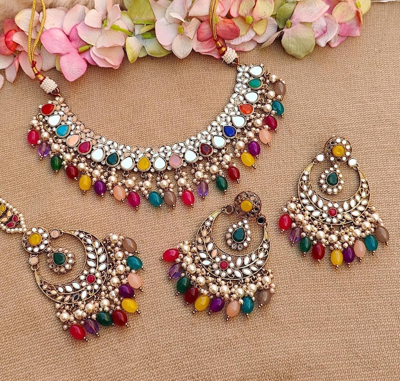 Puede incluir: Un conjunto de collar y pendientes de tono dorado con un dise&ntilde;o multicolor. El collar presenta un gran colgante redondo con una superficie de espejo y est&aacute; adornado con piedras de colores y perlas. Los pendientes tienen forma de media luna y presentan un dise&ntilde;o similar con piedras de colores y perlas.