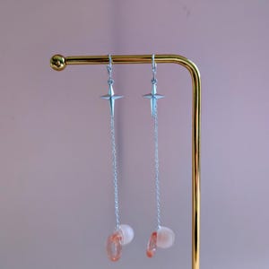 May include: A pair of silver-toned drop earrings featuring a four-pointed star charm attached to a delicate chain. Each earring ends with a small, translucent, pale pink spherical bead and a matching circular ring charm in a soft peach hue.