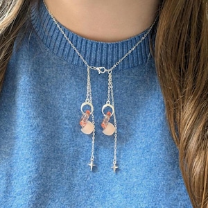 May include: A silver-toned necklace featuring a delicate chain that forms a choker style, with two dangling vertical chains. Each drop includes a small silver ring, a translucent peach-colored bead, and a tiny four-pointed star charm at the end.