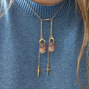 Puede incluir: Un collar tipo lariat en tono dorado con una cadena fina y dos secciones colgantes, cada una adornada con un peque&ntilde;o anillo transl&uacute;cido color melocot&oacute;n, una cuenta esmerilada y un dije de estrella de cuatro puntas en el extremo.